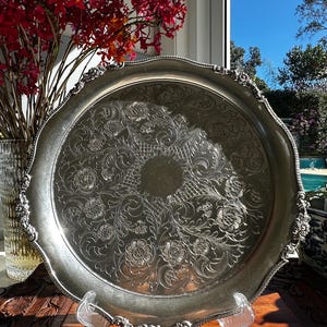 May include: A round silver-toned metal tray featuring an intricate embossed floral pattern across its surface. The tray has a scalloped edge with detailed relief work and a smooth, circular centre, supported by a clear acrylic display stand.