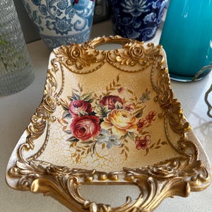 May include: A rectangular decorative tray featuring a cream-coloured centre with a crackled finish and a central floral bouquet of red, yellow, and pink roses. The tray is framed by an ornate, raised gold-tone border with sculpted rose details.