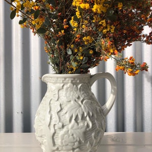 May include: A white ceramic jug with an embossed floral design. The jug is filled with a bouquet of yellow and orange flowers.