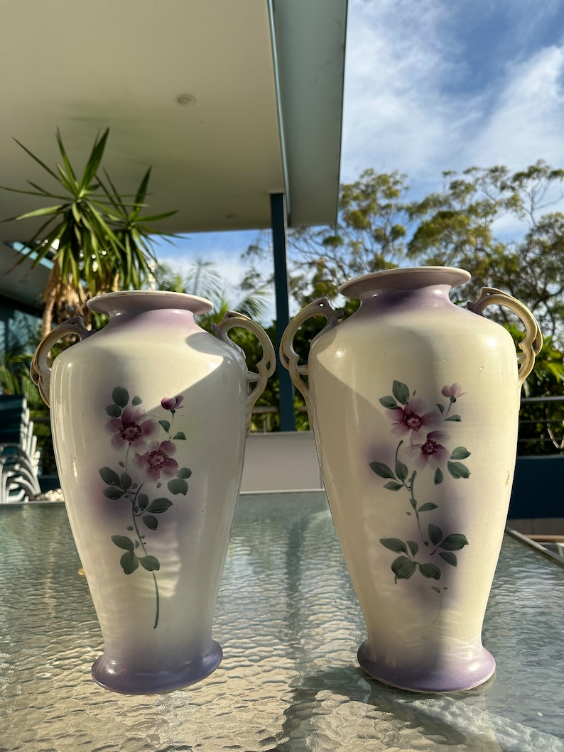 May include: Two matching cream-colored vases with purple accents and floral designs. Each vase has a flared base, a wide opening, and gold-colored handles. The floral design features purple flowers and green leaves.