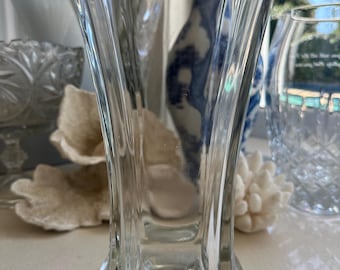 May include: A clear glass vase with a wide mouth and a flared base. The vase has a geometric design with straight lines and a slightly rounded shape.