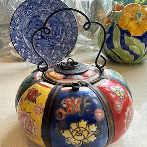 May include: A round, multi-colored ceramic teapot with a metal handle and lid. The teapot is decorated with floral patterns and Chinese characters.