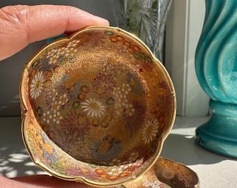 May include: Two small, decorative bowls with a floral pattern. The bowls have a gold rim and a detailed design featuring white, yellow, and red flowers on a gold background. One bowl is held in a hand, and the other sits nearby.