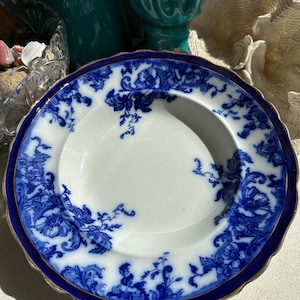 Puede incluir: Un plato blanco con un dise&ntilde;o floral azul y un borde azul oscuro. El plato tiene un borde festoneado con un ribete dorado. El patr&oacute;n floral es intrincado y cubre el borde y parte de la superficie.