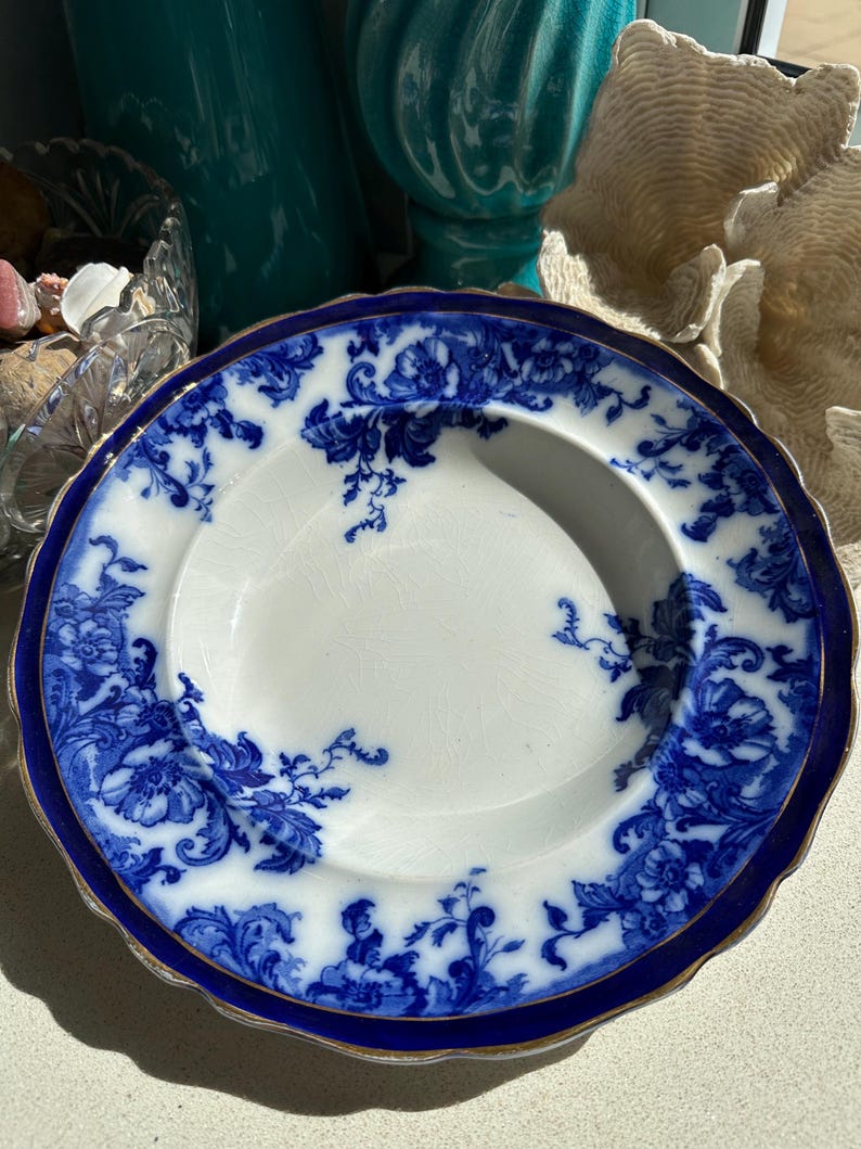 Puede incluir: Un plato blanco con un dise&ntilde;o floral azul y un borde azul oscuro. El plato tiene un borde festoneado con un ribete dorado. El patr&oacute;n floral es intrincado y cubre el borde y parte de la superficie.