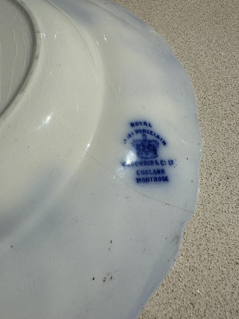 Puede incluir: Un plato de porcelana blanca con un dise&ntilde;o floral azul y un borde festoneado. El plato tiene un sello azul que dice "Royal Porcelain J.A. Wood & Co Ltd England Montrose."