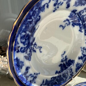 Puede incluir: Un plato de porcelana blanco y azul con un dise&ntilde;o floral. El plato tiene un borde azul oscuro con un ribete dorado. El patr&oacute;n floral est&aacute; en tonos de azul. El plato es parte de un juego de vajilla.