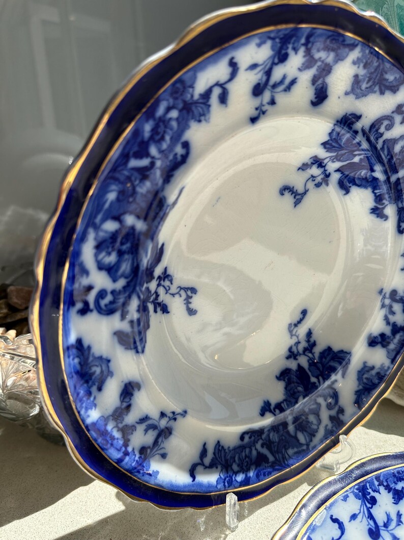 Puede incluir: Un plato de porcelana blanco y azul con un dise&ntilde;o floral. El plato tiene un borde azul oscuro con un ribete dorado. El patr&oacute;n floral est&aacute; en tonos de azul. El plato es parte de un juego de vajilla.