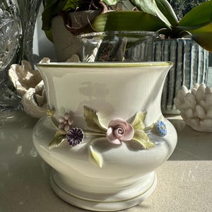 May include: A white ceramic planter with a green rim and raised floral details. The flowers are pink, purple, and blue.