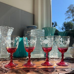 May include: Four stemmed glasses with deep red bases and clear bowls featuring etched white floral patterns and thin gold rims. The glasses are arranged in a row on a dark brown carved wooden tray, showcasing a classic design with a vibrant colour contrast.