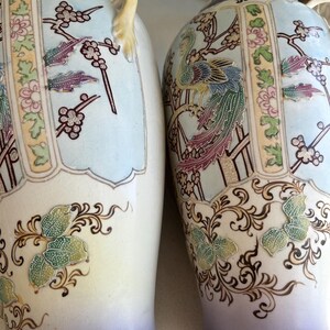 May include: Two decorative vases with a light blue and cream color scheme. The vases feature detailed illustrations of birds, flowers, and butterflies. The handles are a light yellow color. The base of the vases is a light purple color.