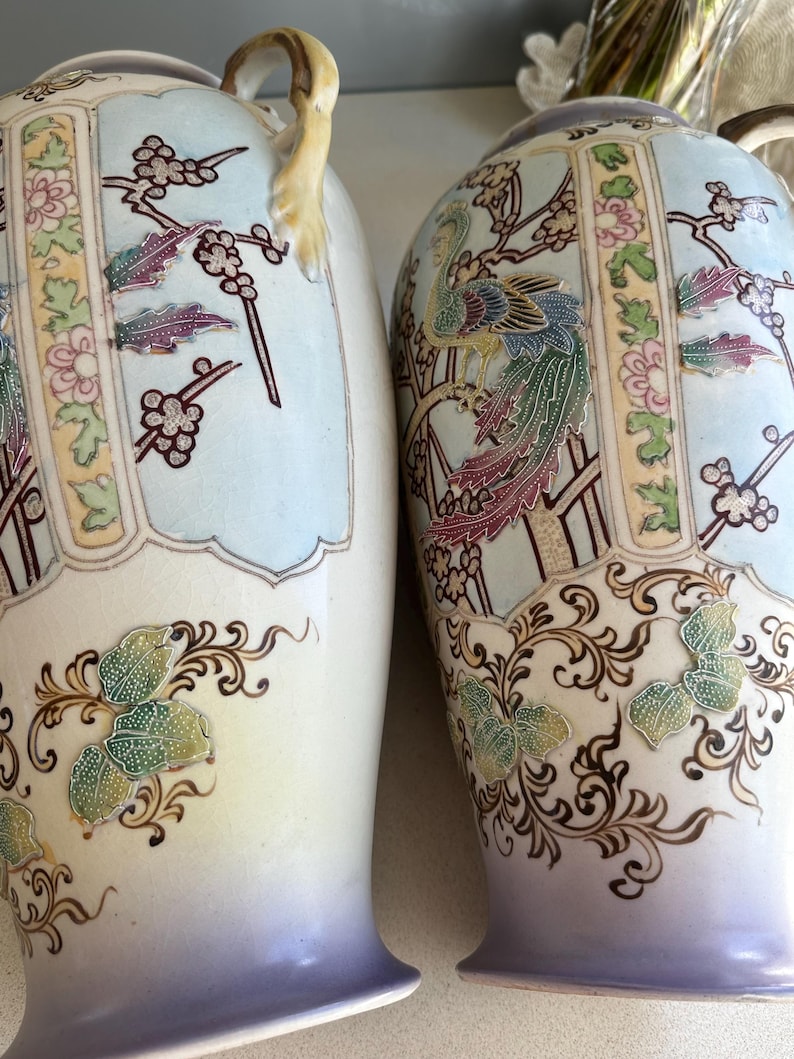 May include: Two decorative vases with a light blue and cream color scheme. The vases feature detailed illustrations of birds, flowers, and butterflies. The handles are a light yellow color. The base of the vases is a light purple color.