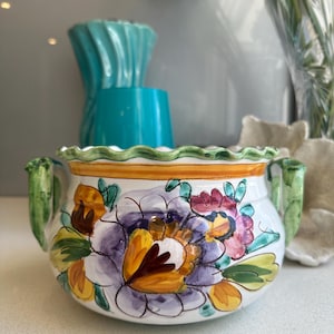 May include: A white ceramic planter with a floral design in purple, yellow, and pink. The planter has two green handles and a scalloped rim. A yellow band encircles the top. Other turquoise vases and flowers are in the background.