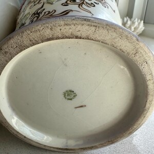 May include: A close-up of the base of a decorative ceramic vase. The vase has a cream-colored base with a light blue and white patterned design. The base has a textured, aged appearance.