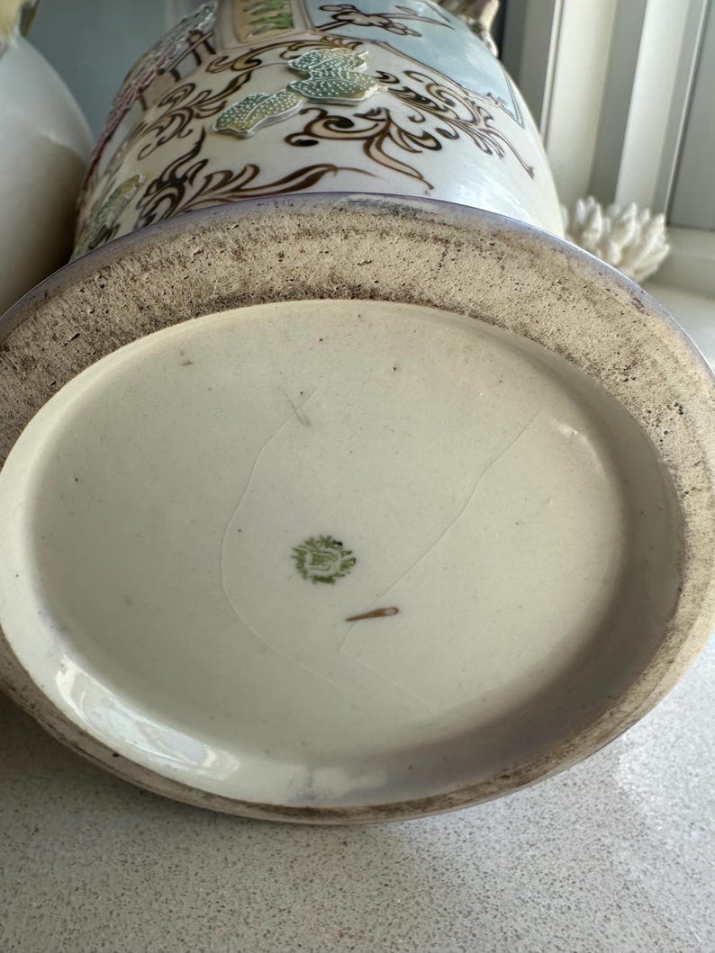 May include: A close-up of the base of a decorative ceramic vase. The vase has a cream-colored base with a light blue and white patterned design. The base has a textured, aged appearance.