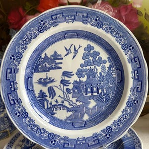 May include: A white and blue ceramic plate with a Willow pattern design. The plate features a detailed landscape scene with pagodas, bridges, and birds. The rim has a decorative border. This plate is part of a dinnerware set.