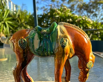 May include: A ceramic horse figurine with a brown, green, and yellow glaze. The horse is in a standing position with its head lowered. The saddle is green and the figurine has decorative details.