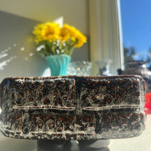 Könnte beinhalten: Ein rechteckiger, brauner Keramik-Blumentopf mit strukturierter Oberfläche. Der Pflanztopf hat einen dunkelbraunen Sockel und steht auf einer weißen Oberfläche. Gelbe Blumen in einer türkisfarbenen Vase sind im Hintergrund zu sehen.