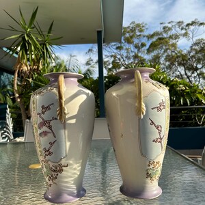 May include: Two matching antique vases. Each vase has a cream-colored body with a lavender base and rim. The vases feature a floral design with gold accents and a decorative handle. The vases are displayed on a glass table.