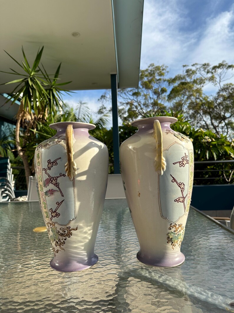 May include: Two matching antique vases. Each vase has a cream-colored body with a lavender base and rim. The vases feature a floral design with gold accents and a decorative handle. The vases are displayed on a glass table.