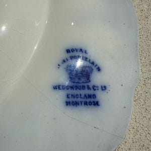 Puede incluir: Un plato de porcelana blanco y azul con borde festoneado. El plato presenta un sello azul que dice "ROYAL PORCELAIN", "WEDGWOOD & CO LTD", "ENGLAND" y "MONTROSE".