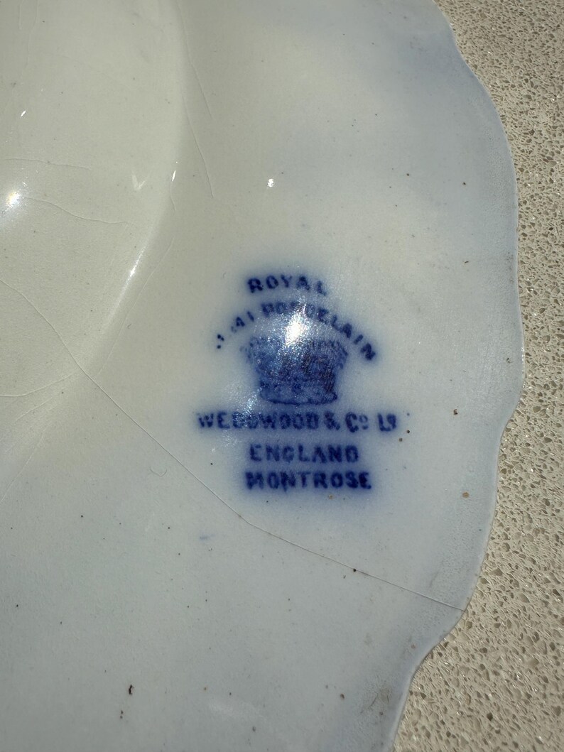 Puede incluir: Un plato de porcelana blanco y azul con borde festoneado. El plato presenta un sello azul que dice "ROYAL PORCELAIN", "WEDGWOOD & CO LTD", "ENGLAND" y "MONTROSE".