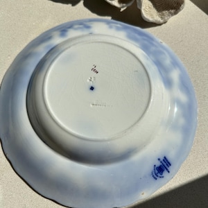 Puede incluir: Plato de cer&aacute;mica vintage con un dise&ntilde;o floral azul claro y blanco. La parte inferior del plato presenta un &aacute;rea circular central y un sello azul con texto. El borde tiene un borde festoneado.