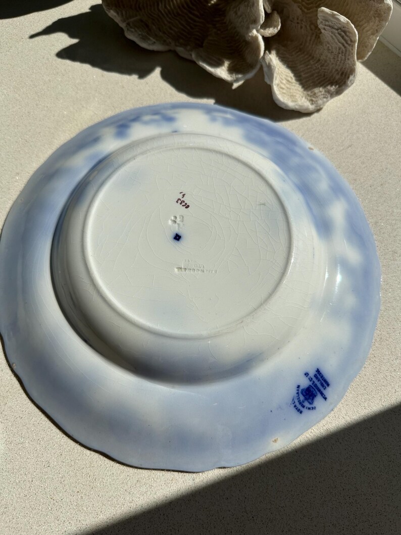 Puede incluir: Plato de cer&aacute;mica vintage con un dise&ntilde;o floral azul claro y blanco. La parte inferior del plato presenta un &aacute;rea circular central y un sello azul con texto. El borde tiene un borde festoneado.