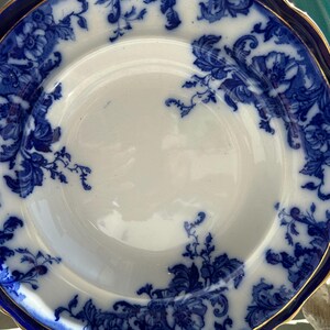Puede incluir: Plato blanco con dise&ntilde;o floral azul y borde dorado. El plato tiene un borde festoneado y un borde azul oscuro. El patr&oacute;n floral es intrincado y cubre el borde y parte de la superficie del plato.