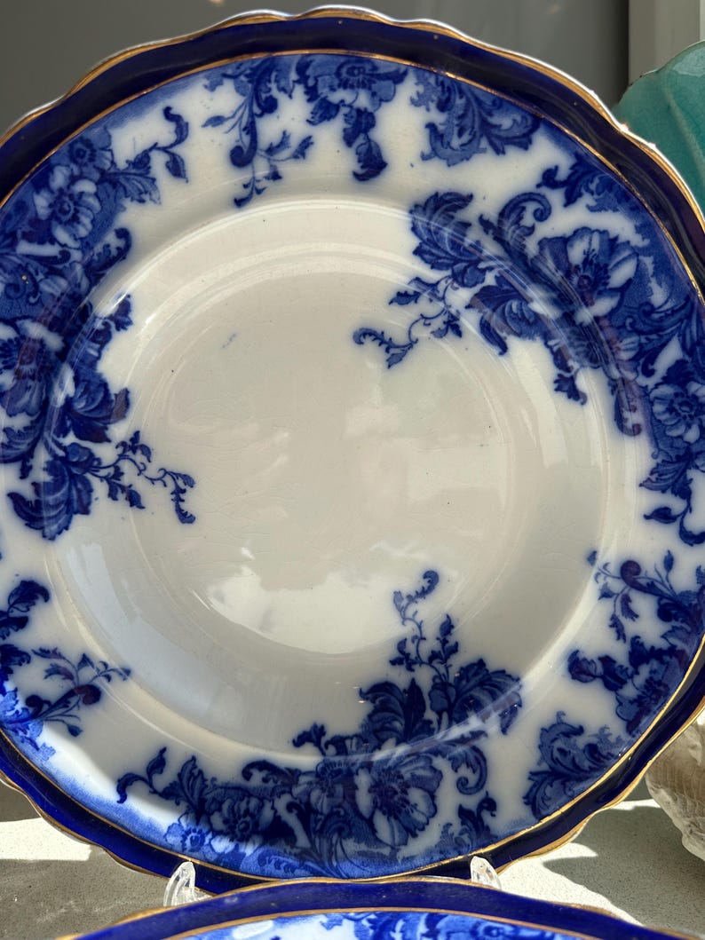 Puede incluir: Plato blanco con dise&ntilde;o floral azul y borde dorado. El plato tiene un borde festoneado y un borde azul oscuro. El patr&oacute;n floral es intrincado y cubre el borde y parte de la superficie del plato.