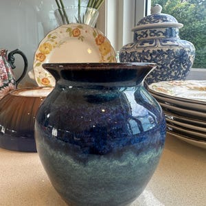 May include: A blue and green ceramic vase with a brown rim. The vase is round and has a wide opening. It is sitting on a white surface.