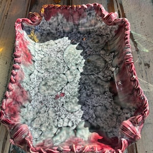 Könnte beinhalten: Eine Servierplatte aus Keramik mit einem gewellten Rand. Die Platte ist mit einer craquelé-Glasur in den Farben Rosa, Grau und Weiß verziert.