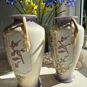 May include: Two cream-colored ceramic vases with a lavender rim and base. Each vase features a decorative panel with floral and bird motifs in shades of pink, green, and gold. The vases have curved handles and are displayed in front of a window.
