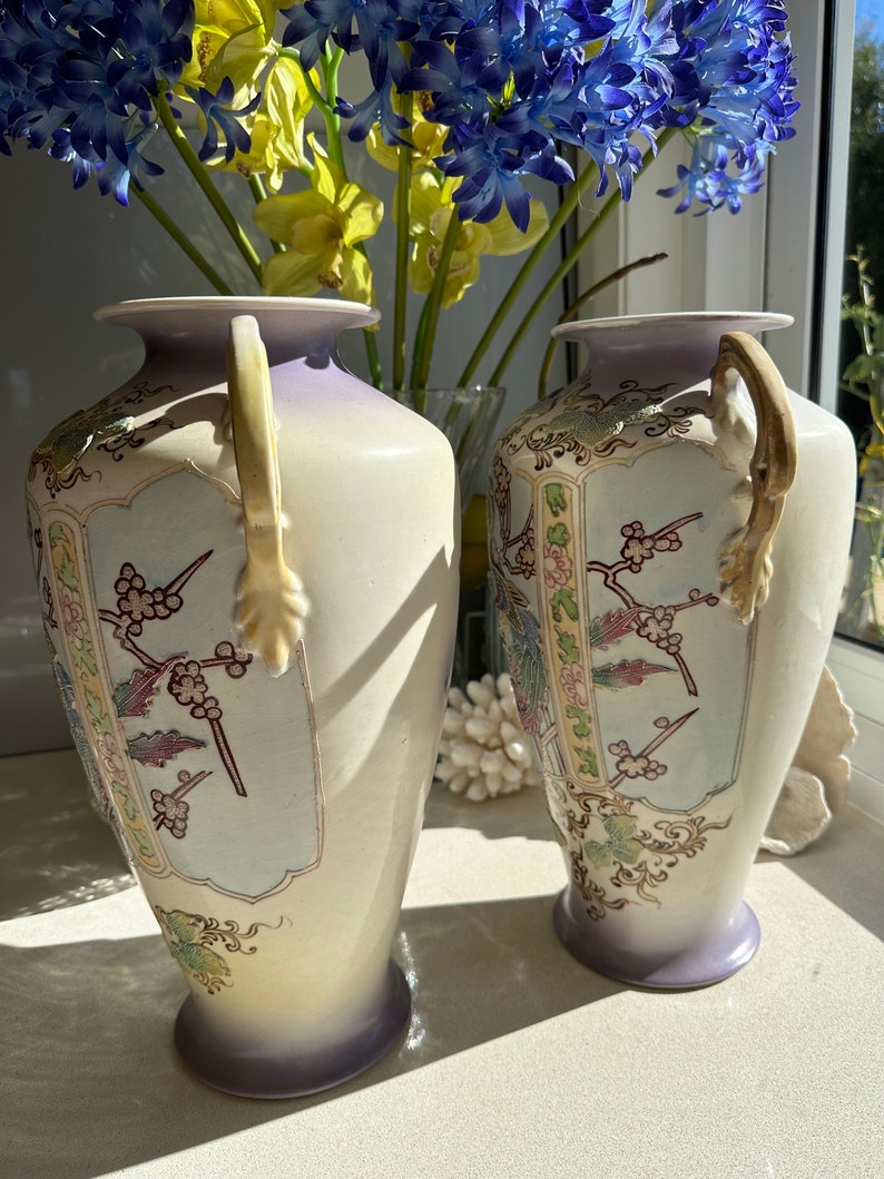 May include: Two cream-colored ceramic vases with a lavender rim and base. Each vase features a decorative panel with floral and bird motifs in shades of pink, green, and gold. The vases have curved handles and are displayed in front of a window.