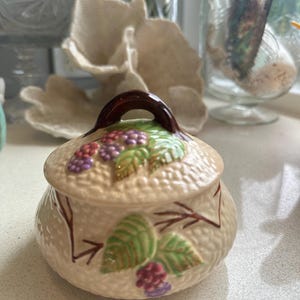 Op de afbeelding: Een keramische pot met een deksel, met een bruin handvat en een ontwerp van groene bladeren en paarse bessen. De pot heeft een getextureerd oppervlak en een crèmekleurige achtergrond.