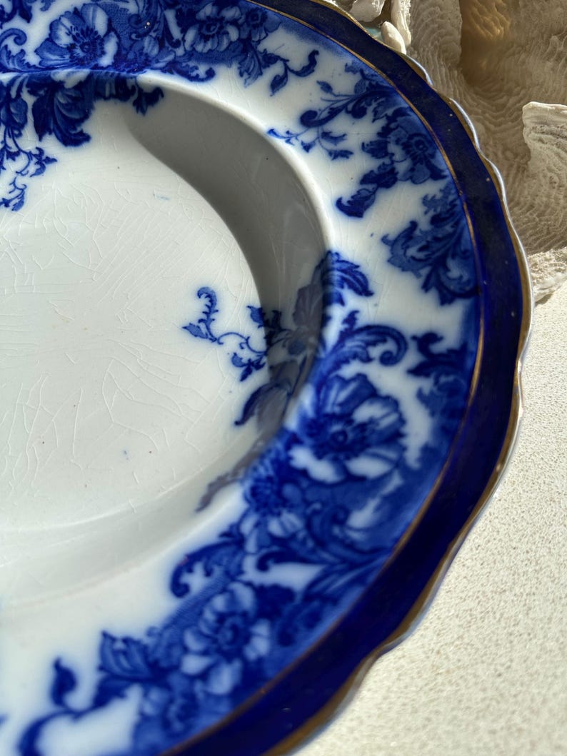Puede incluir: Un plato antiguo blanco y azul con un dise&ntilde;o floral. El plato tiene un borde azul oscuro con un ribete dorado. El dise&ntilde;o floral es detallado, con un borde festoneado. Di&aacute;metro aproximado de 20 cm.