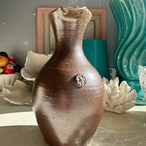 May include: A brown ceramic vase with a textured surface and a decorative floral emblem. The vase has a wide, rounded body and a narrow, fluted neck. The vase is approximately 12 inches tall.