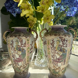 May include: Pair of antique ceramic vases with a light blue and lavender color scheme. Each vase features a detailed peacock design with gold handles and floral accents. The vases are displayed in front of a window with colorful flowers.