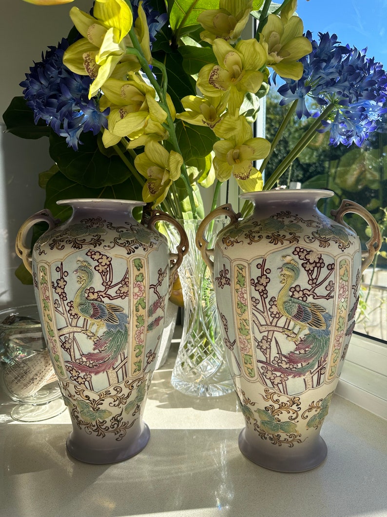 May include: Pair of antique ceramic vases with a light blue and lavender color scheme. Each vase features a detailed peacock design with gold handles and floral accents. The vases are displayed in front of a window with colorful flowers.