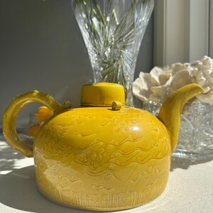 May include: A yellow ceramic teapot with a detailed design of stylised clouds and dragons. The teapot has a round body and a small spout.