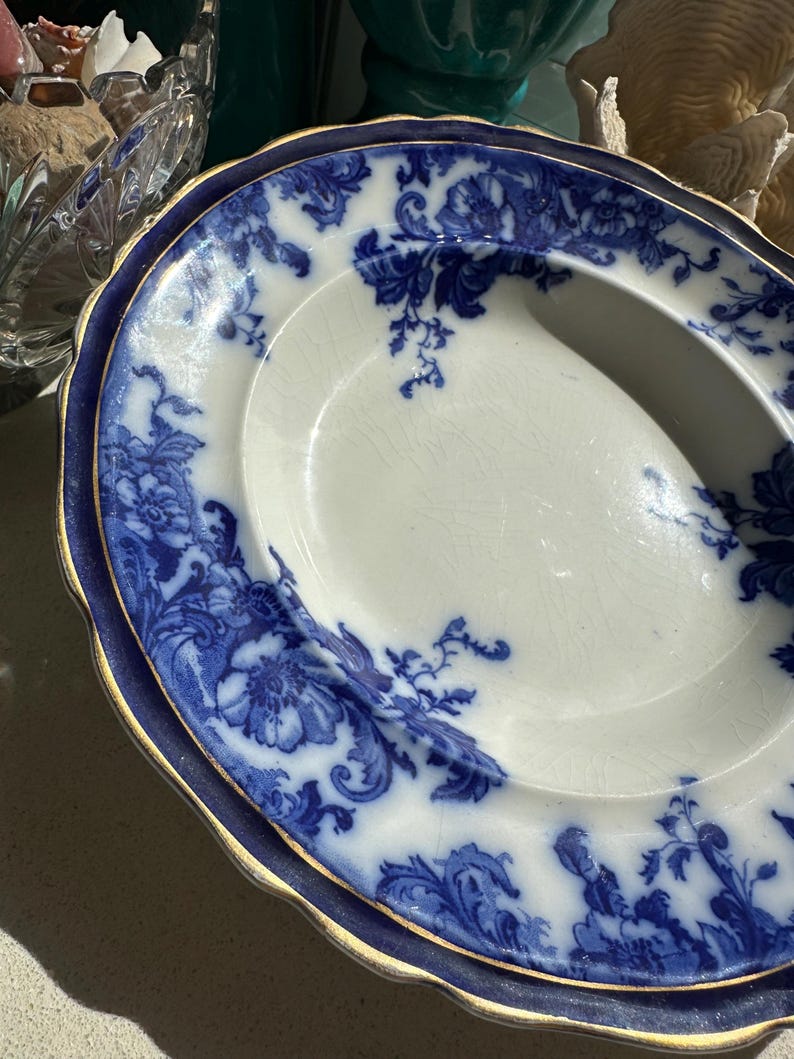 Puede incluir: Plato de servir ovalado antiguo con interior blanco y un patr&oacute;n floral azul. El borde es azul oscuro con un ribete dorado. El plato tiene un asa en un lado y probablemente sea de porcelana.