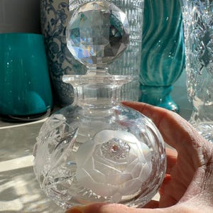 Puede incluir: Un frasco de perfume de vidrio transparente con un tapón esférico facetado. El cuerpo redondo presenta un diseño floral grabado con pétalos de rosa y hojas esmeriladas. El vidrio es transparente y refleja la luz con patrones tallados.