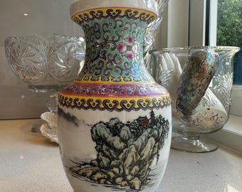 May include: A white ceramic vase with a detailed landscape painting of mountains and a building. The vase has a colorful floral and geometric design around the neck and base. The vase is a decorative item.