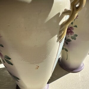 May include: Two off-white ceramic vases with floral designs and lavender bases. The vases have decorative handles and are adorned with hand-painted purple flowers and green leaves. The vases are tall and elegant, suitable for displaying flowers or as decorative pieces.