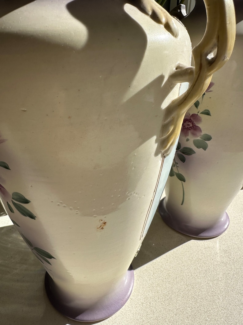 May include: Two off-white ceramic vases with floral designs and lavender bases. The vases have decorative handles and are adorned with hand-painted purple flowers and green leaves. The vases are tall and elegant, suitable for displaying flowers or as decorative pieces.