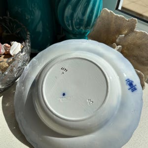 Puede incluir: Un plato de cer&aacute;mica blanca con borde festoneado, con un dise&ntilde;o floral azul. La parte inferior muestra marcas, incluido el texto "Royal Winton, England". El plato es probablemente vintage y adecuado para exhibir o cenar.