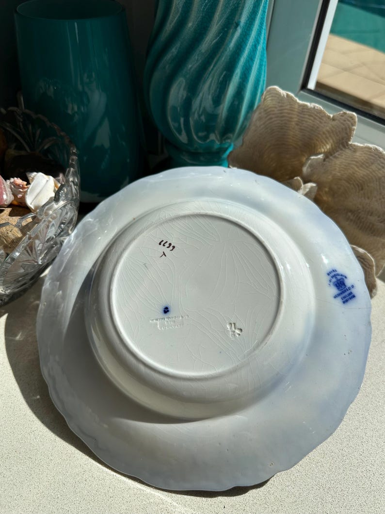 Puede incluir: Un plato de cer&aacute;mica blanca con borde festoneado, con un dise&ntilde;o floral azul. La parte inferior muestra marcas, incluido el texto "Royal Winton, England". El plato es probablemente vintage y adecuado para exhibir o cenar.