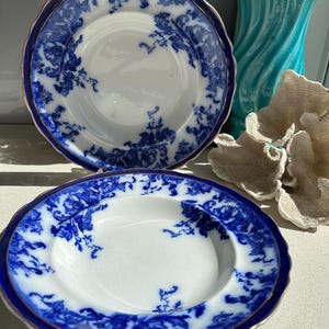 Puede incluir: Dos platos antiguos de porcelana blanca con un dise&ntilde;o floral azul y un borde dorado. Los platos tienen un borde festoneado y un borde azul oscuro. Un plato es ligeramente m&aacute;s hondo que el otro. Los platos son probablemente para uso decorativo o para servir.