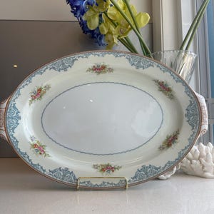 May include: An oval porcelain serving platter featuring a cream-coloured centre and a decorative border. The rim is adorned with intricate light blue scrollwork patterns and small, colourful floral clusters in shades of pink, yellow, and green.