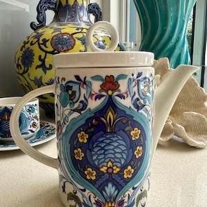 May include: A ceramic teapot with a lid, adorned with a detailed blue, teal, yellow, and red floral design. The teapot has a white handle and spout. Other decorative vases are in the background.